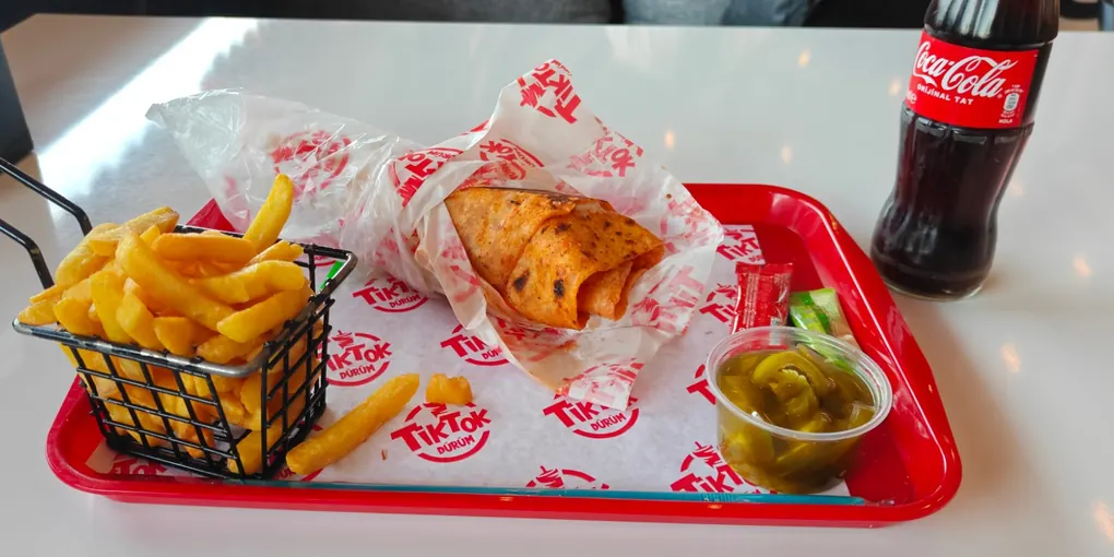 A dürüm in branded paper on a red tray, fries in a wire basket and a bottle of cola